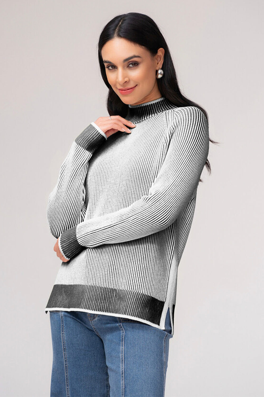 A woman modeling a ribbed knit mock neck sweater with a high-low hem and contrasting black bands, paired with blue jeans against a light gray studio background.