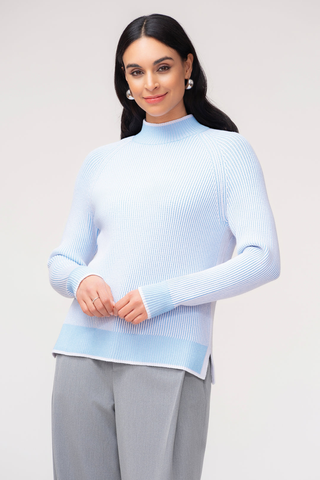 A woman modeling a light blue ribbed mock-neck sweater with a contrasting light blue band at the hem, paired with gray trousers, against a plain light gray studio background.