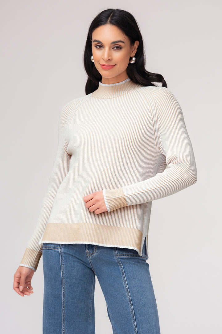 A woman modeling a ribbed knit mock neck sweater in cream with a beige band at the hem and cuffs, paired with blue jeans, against a light gray studio background.