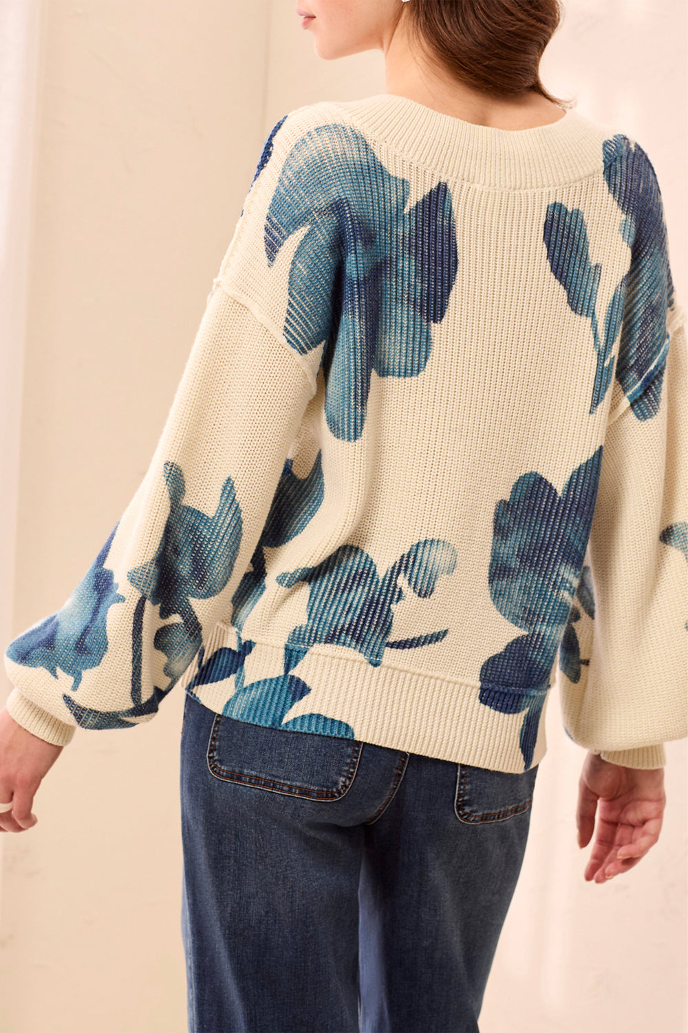A woman shown from the back wearing a cream sweater with a blue floral pattern and jeans in front of a beige background. 