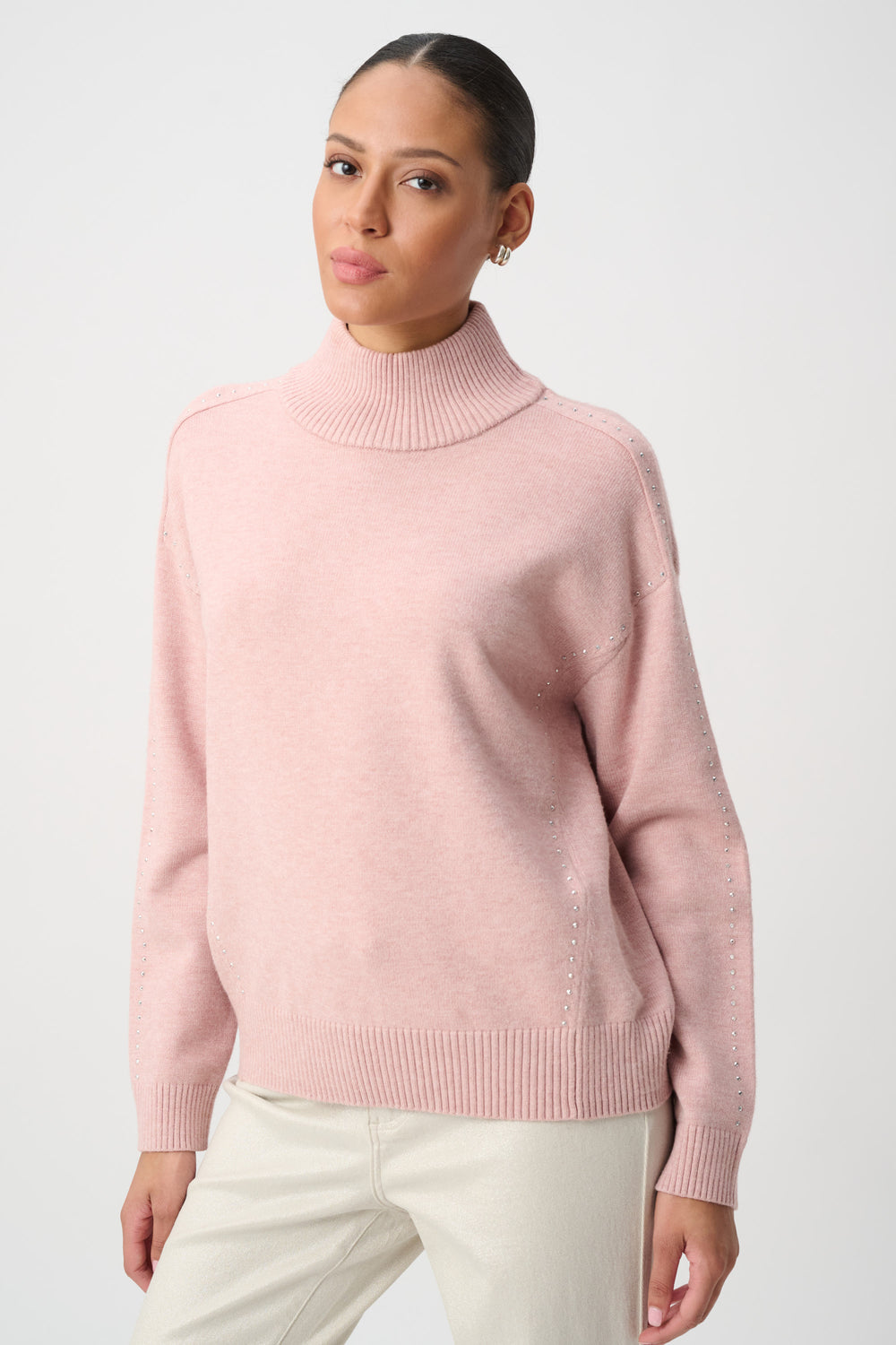 A woman wearing a light pink ribbed turtleneck sweater and cream trousers, photographed against a clean light gray studio background.