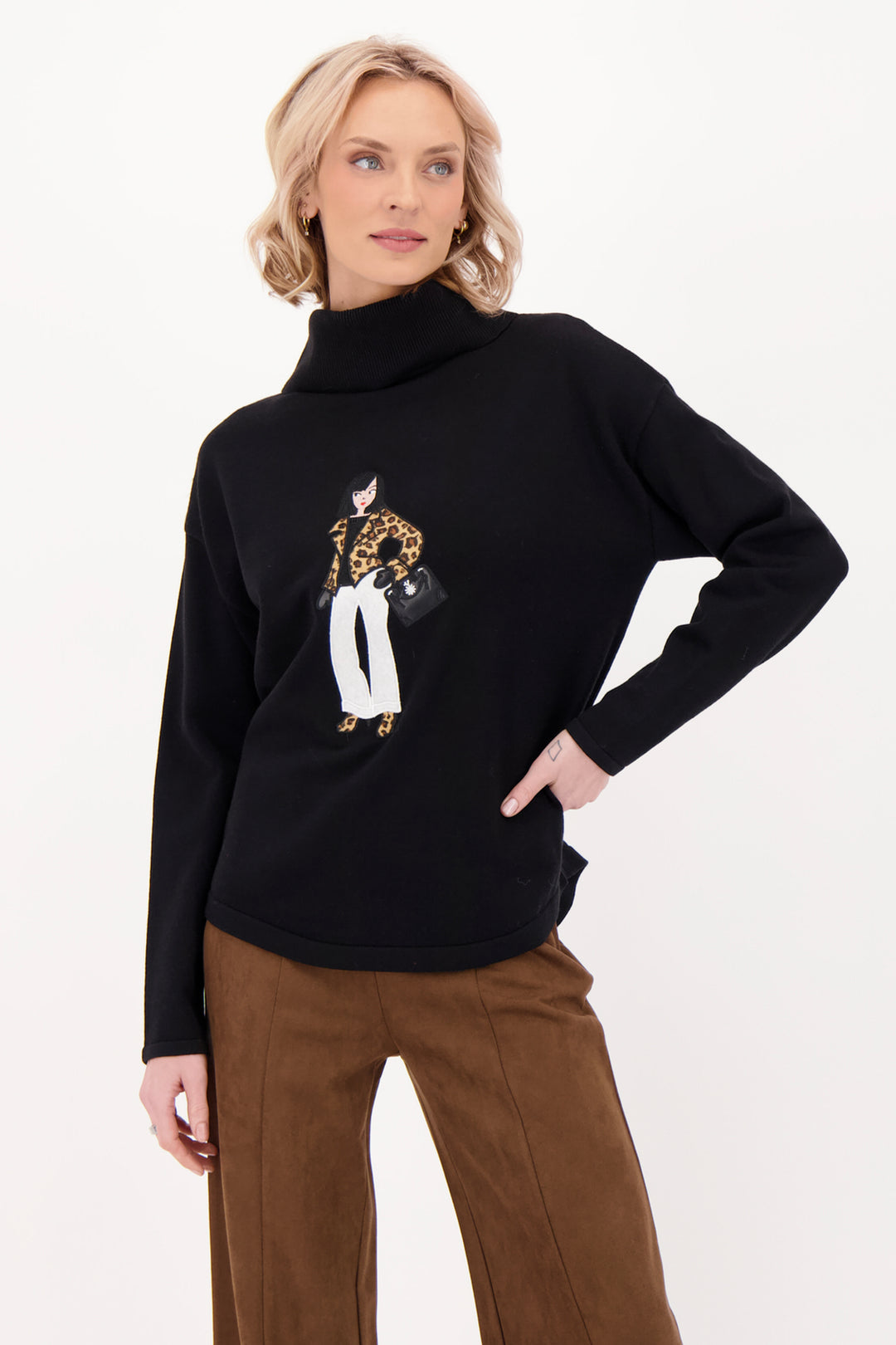 A woman modelling a black sweater with a girl on the front with brown suede pants in front of a grey background. 