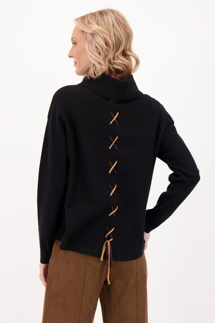 A woman modelling a black sweater with a girl on the front with brown suede pants in front of a grey background. 