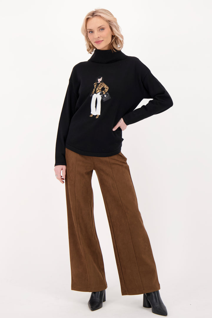 A woman modelling a black sweater with a girl on the front with brown suede pants in front of a grey background. 