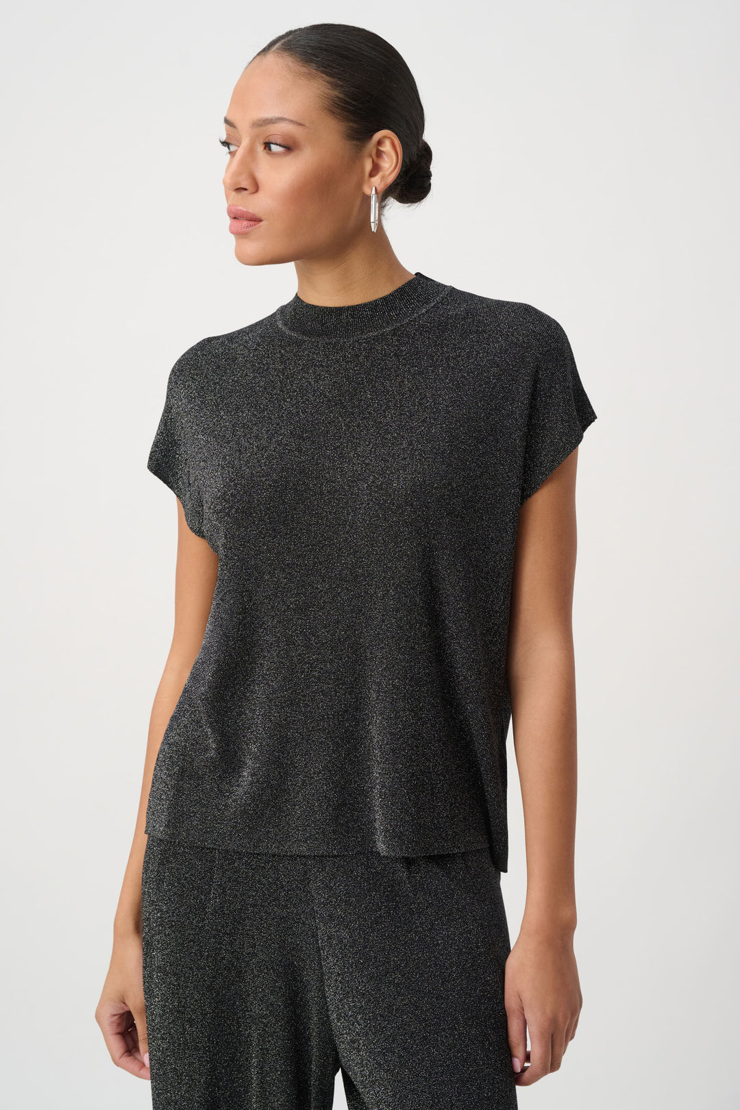 A woman modelling a black shimmery t-shirt top with a mock neck and matching pants standing in front of a light grey background. 
