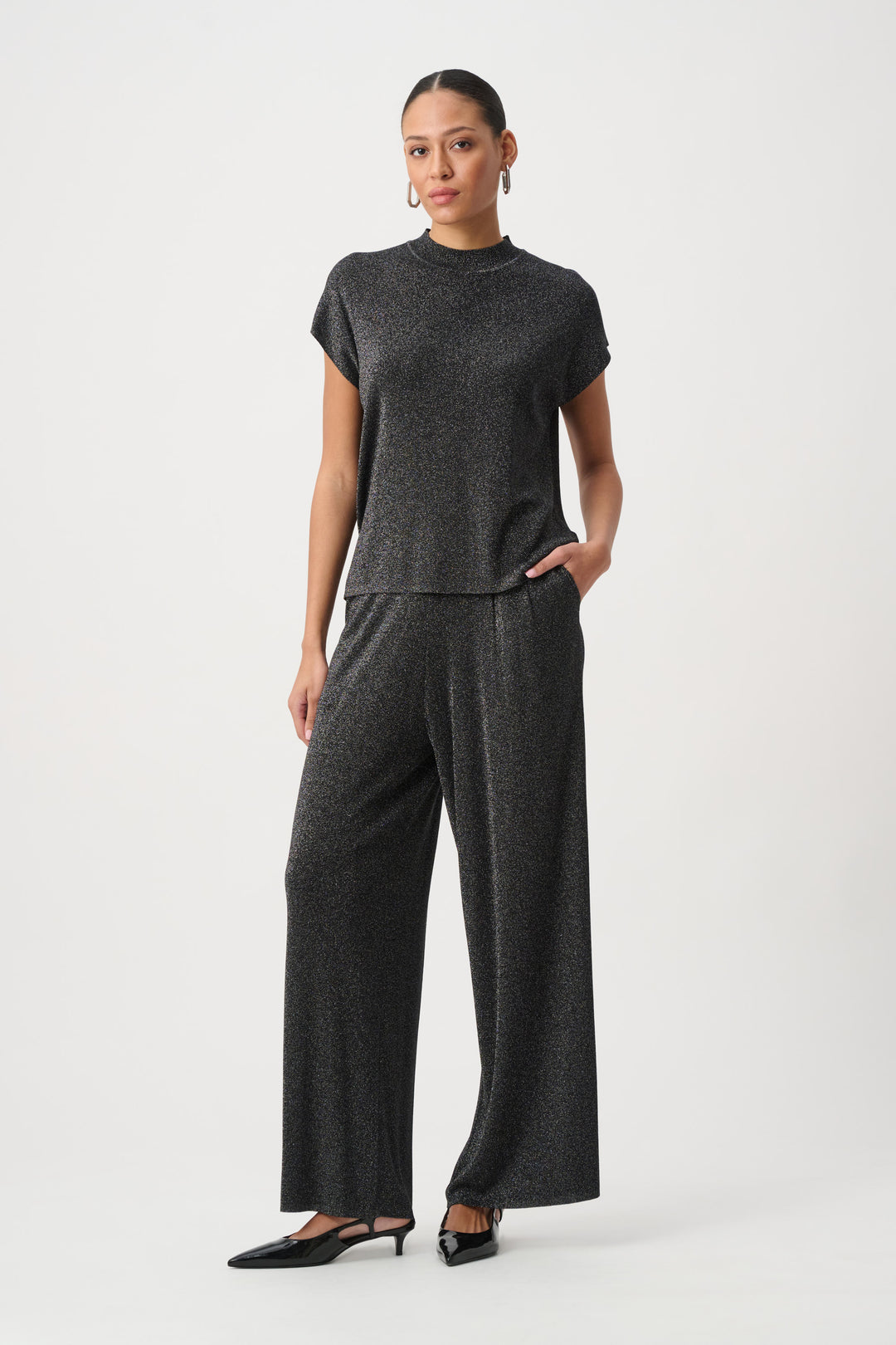 A woman modelling a black shimmery t-shirt top with a mock neck and matching pants standing in front of a light grey background. 