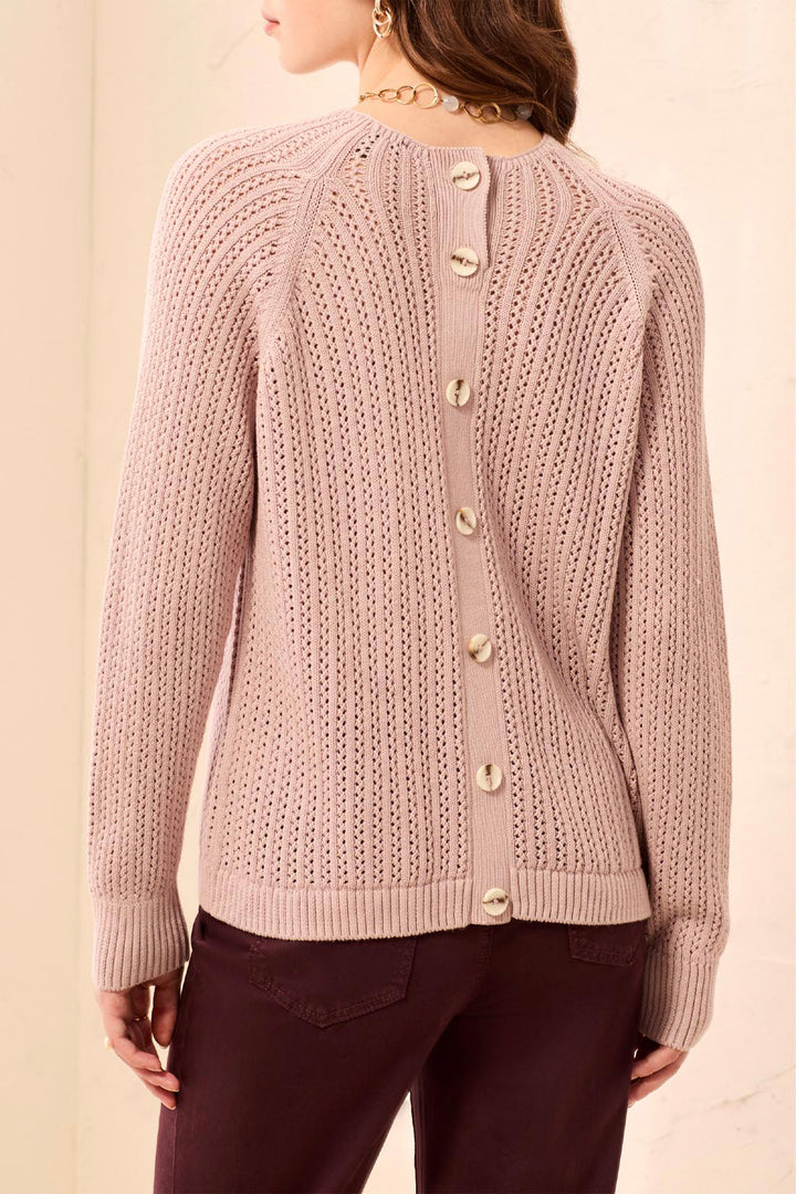 A woman shown from the back modelling a blush pink, loose knit long sleeve sweater with buttons up the back standing in front of a light beige background. 