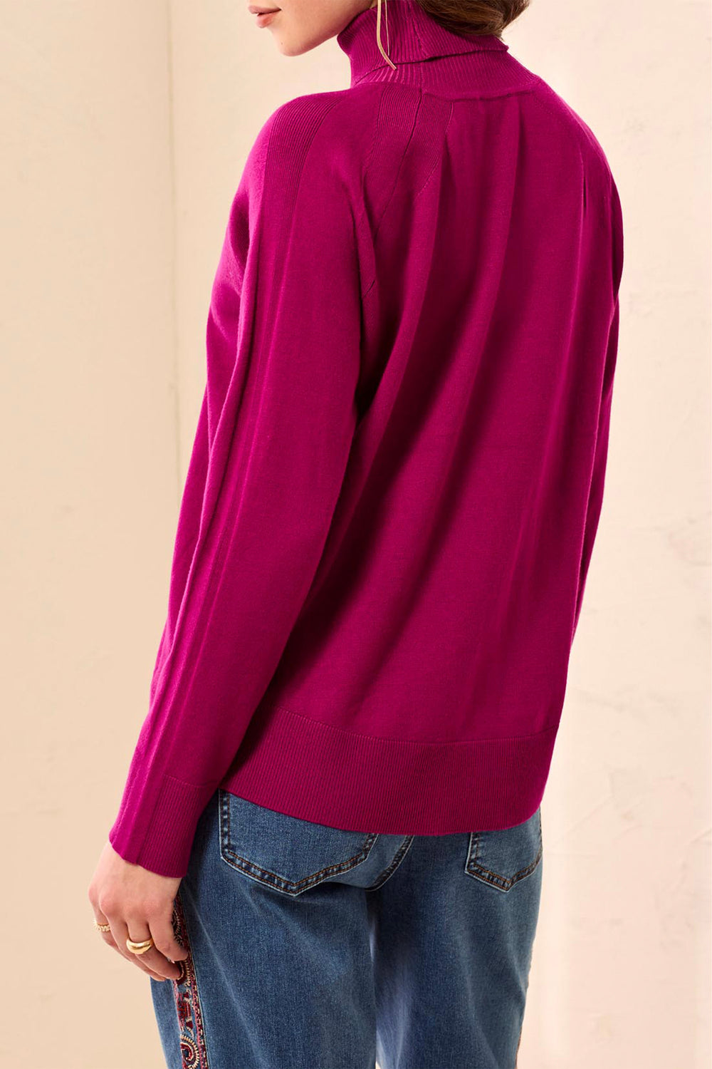 A woman shown from the back modelling a long sleeve magenta turtleneck sweater and jeans standing in front of a light beige background. 