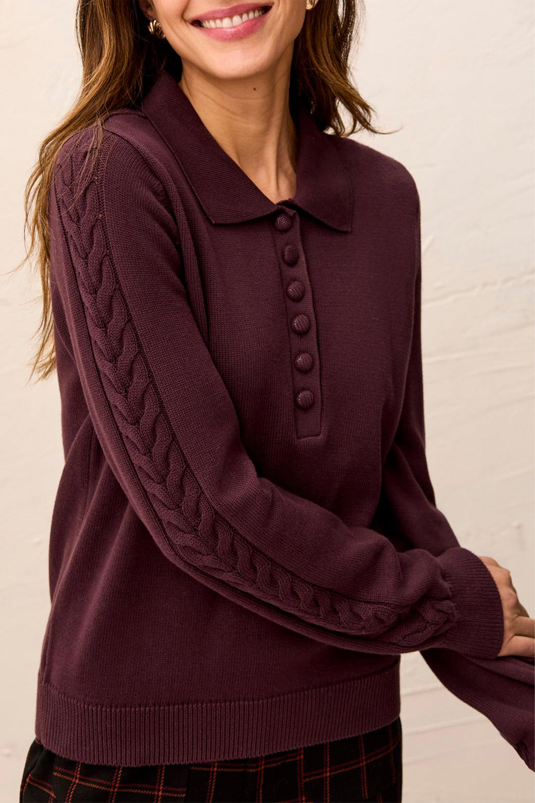 A woman modelling a brown polo style long sleeve sweater with cable knit details up the sleeves. She is standing in front of a light beige background. 