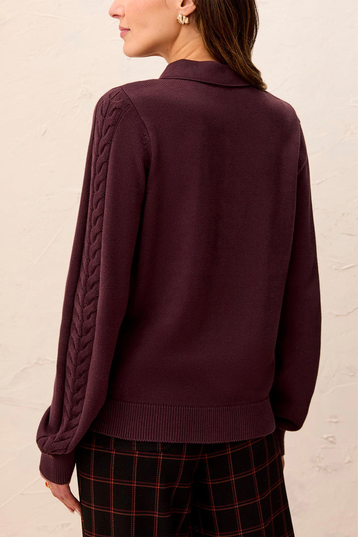 A woman shown from the back modelling a polo style long sleeve sweater with cable knit details up the sleeves. She is standing in front of a light beige background. 