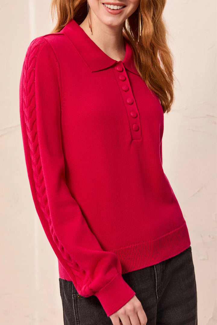 A woman modelling a red polo style long sleeve sweater with cable knit details up the sleeves. She is standing in front of a light beige background. 