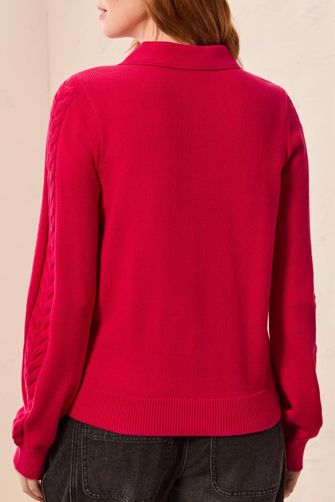 A woman shown from the back modelling a polo style long sleeve sweater with cable knit details up the sleeves. She is standing in front of a light beige background. 