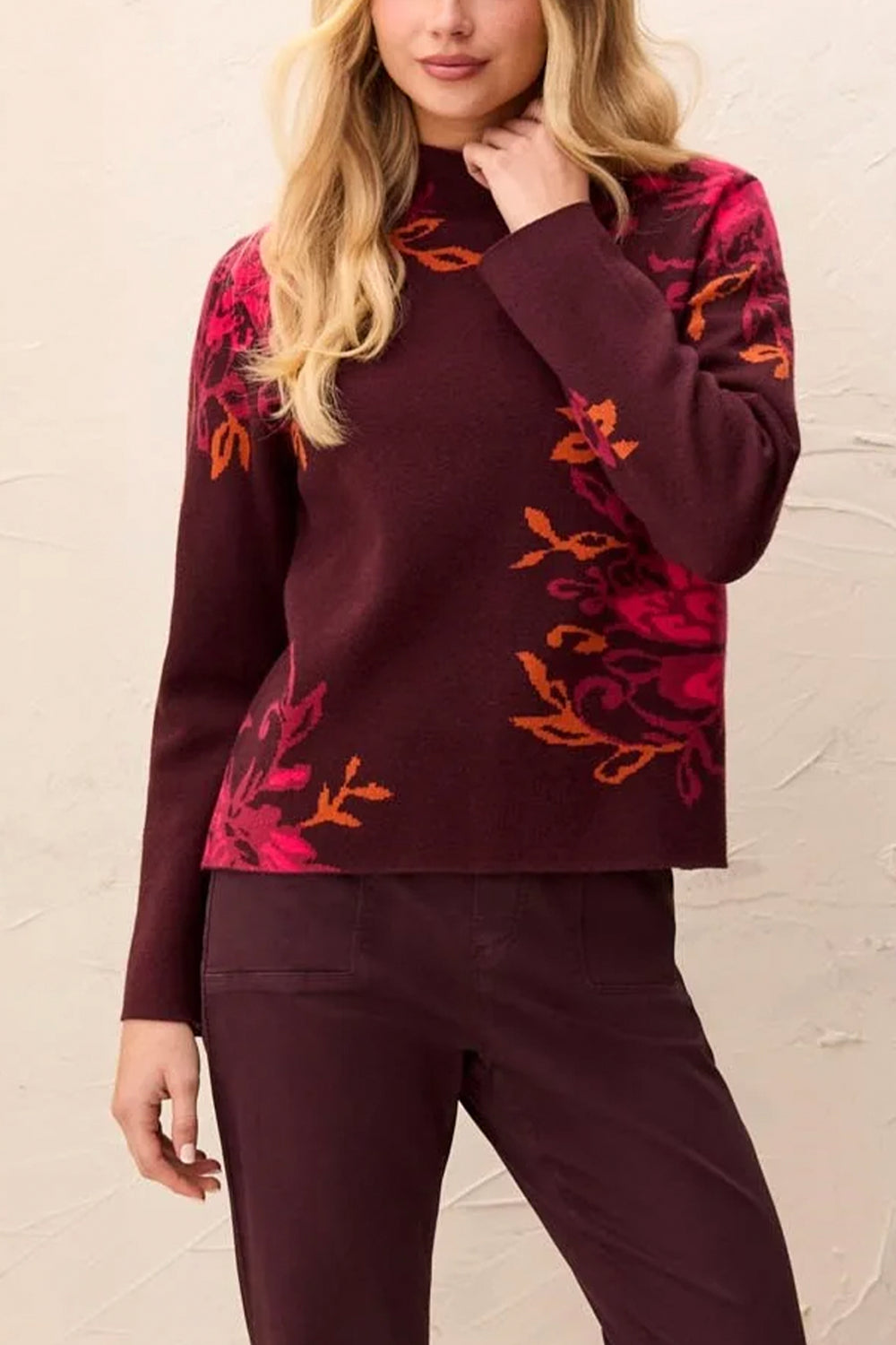 A woman modelling a deep brown long sleeve sweater, with a mock neck and floral details standing in front of a light beige background. 