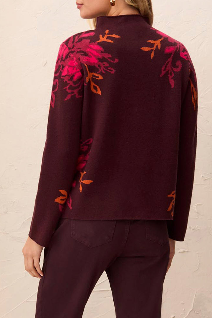 A woman shown from the back modelling a deep brown long sleeve sweater, with a mock neck and floral details standing in front of a light beige background. 