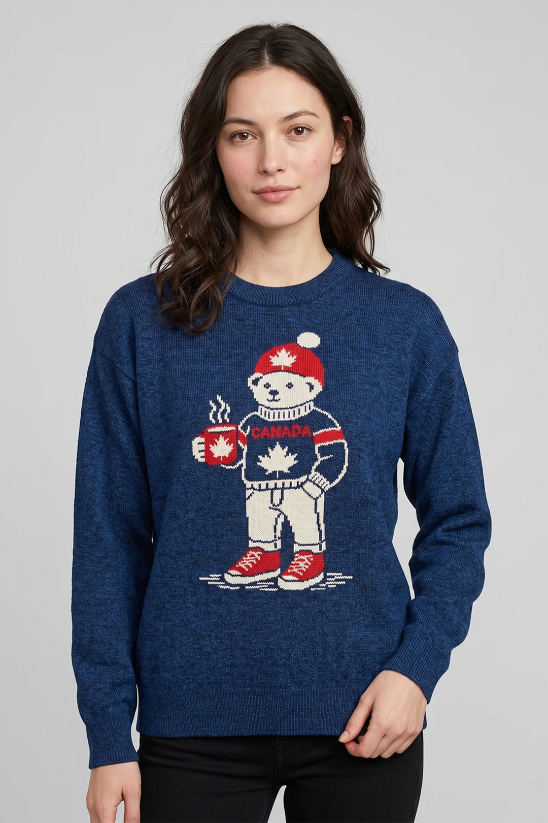 A woman wearing a navy long sleeve sweater with a cute polar bear graphic on the front. The polar bear is holding a steaming maple leaf mug and has a navy Canada sweater on as well as a red maple leaf toque. 