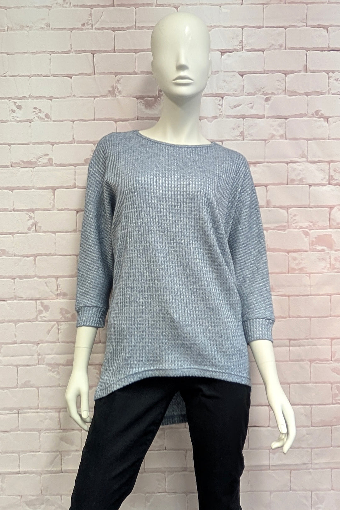 A mannequin wearing a blue metallic high low sweater standing in front of a light brick wall. 