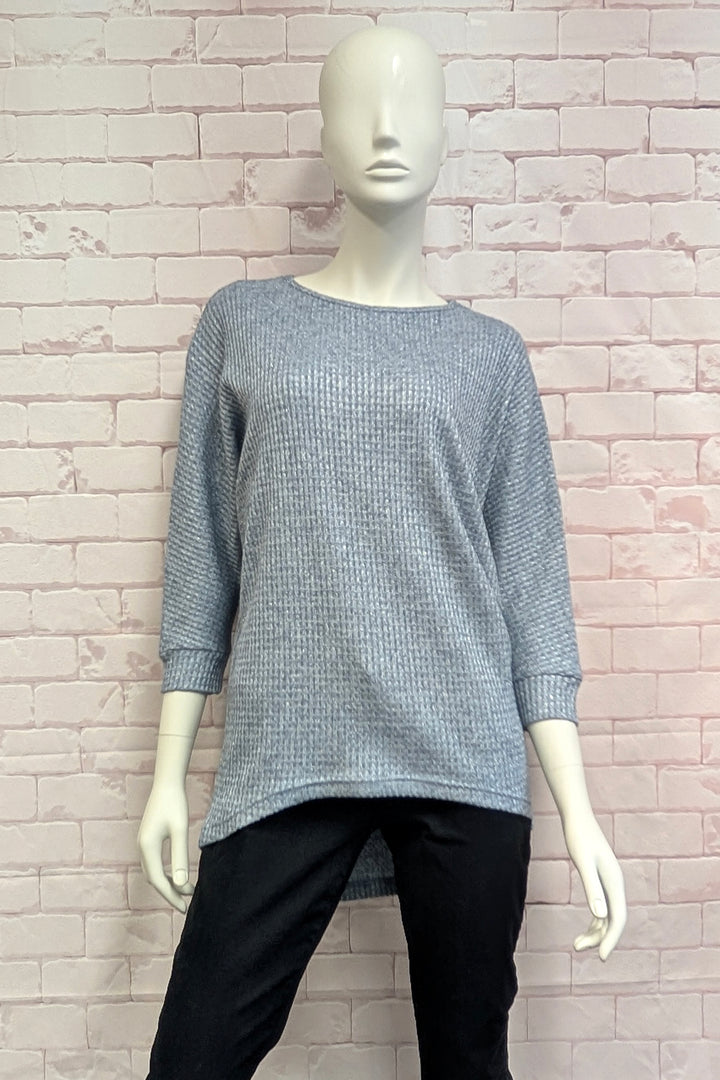 A mannequin wearing a blue metallic high low sweater standing in front of a light brick wall. 
