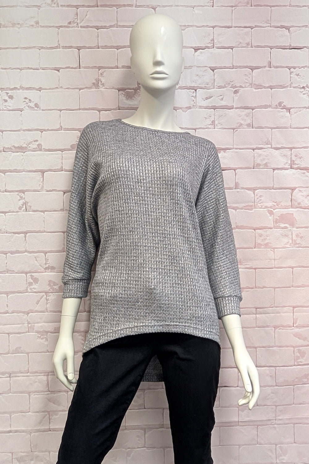A mannequin wearing a grey  metallic high low sweater standing in front of a light brick wall. 