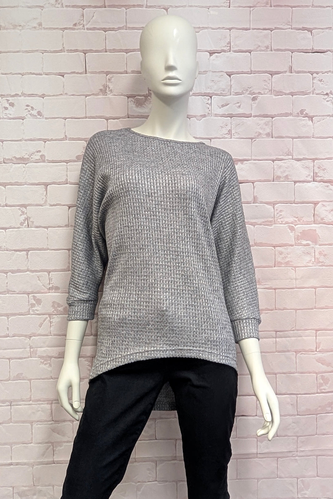 A mannequin wearing a grey  metallic high low sweater standing in front of a light brick wall. 