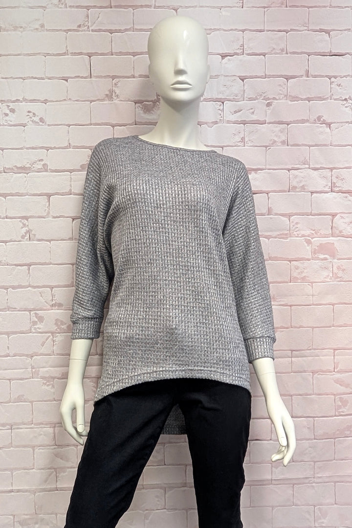 A mannequin wearing a grey  metallic high low sweater standing in front of a light brick wall. 
