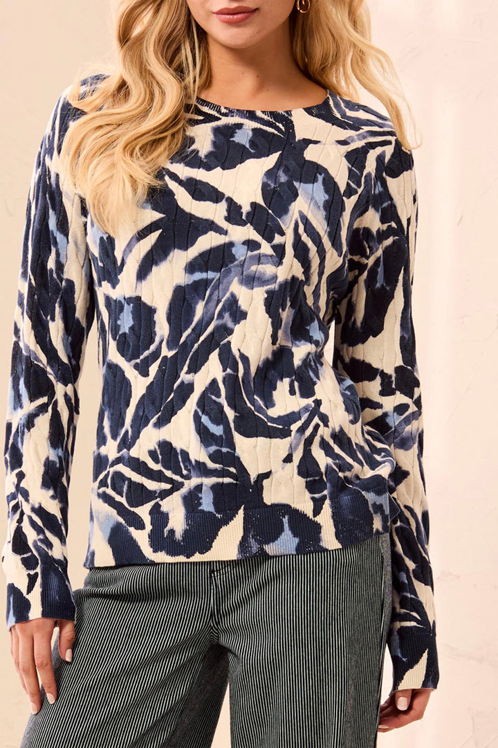 A woman wearing a beige sweater with navy and blue floral details standing in front of a beige background. 