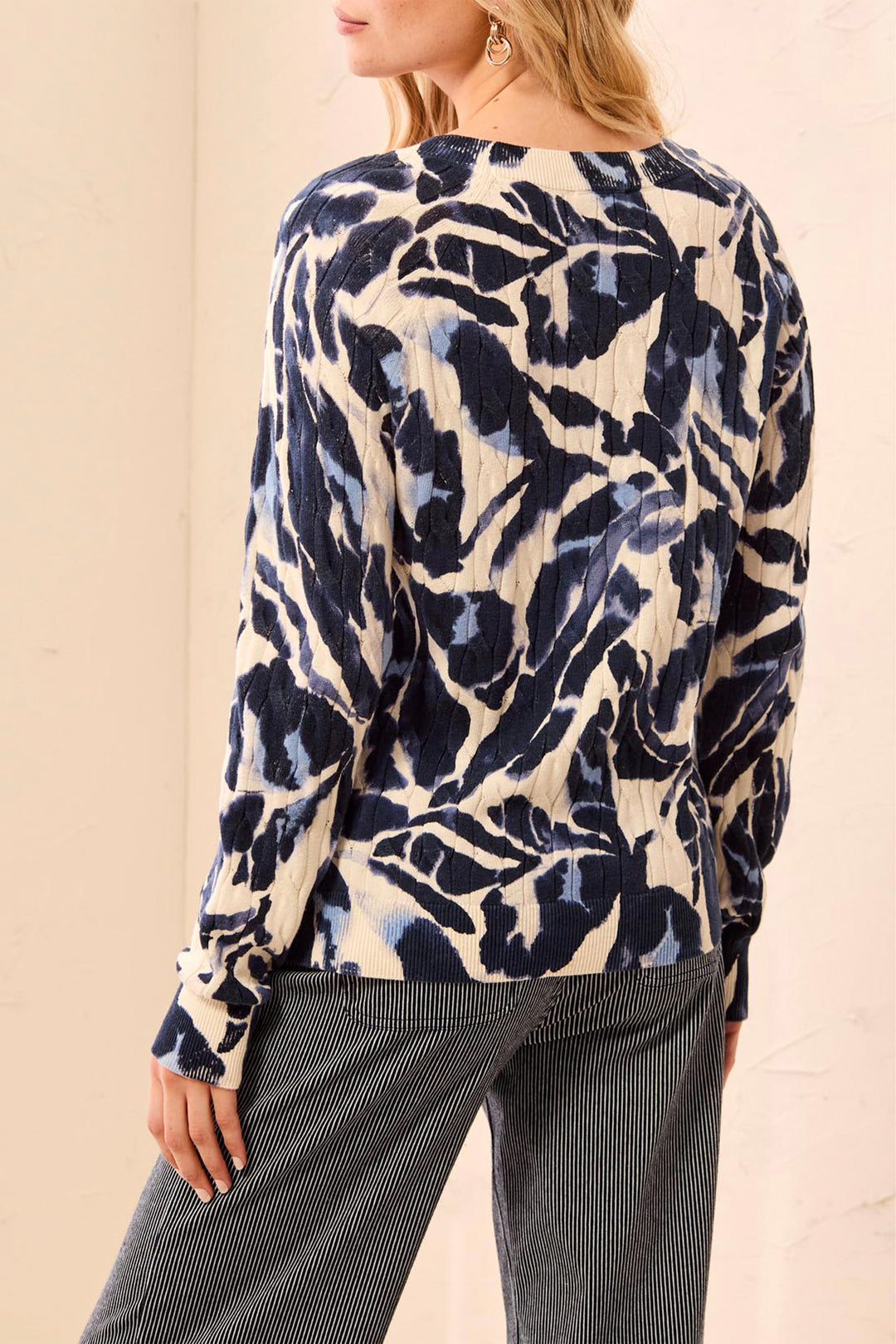 A woman shown from the back wearing a beige sweater with navy and blue floral details standing in front of a beige background. 