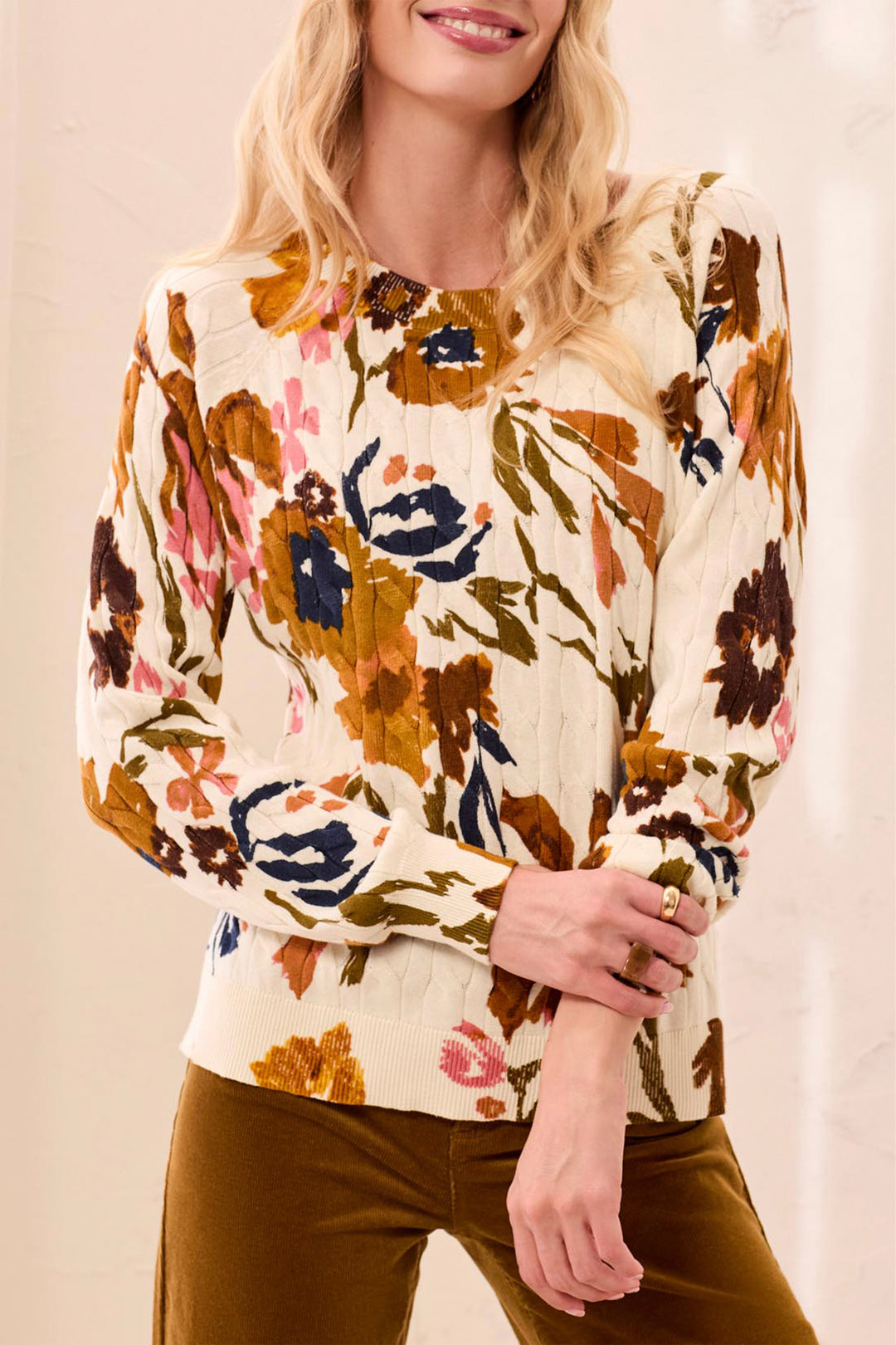 A woman wearing a beige sweater with tan and pink floral details standing in front of a beige background. 