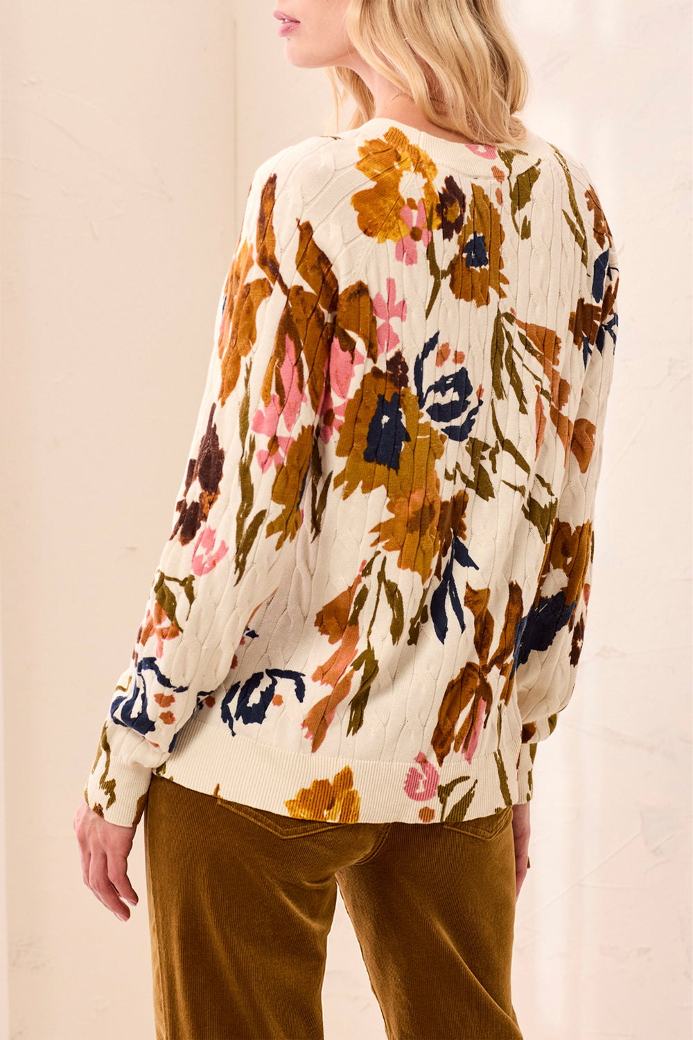 A woman shown from the back wearing a beige sweater with tan and pink floral details standing in front of a beige background. 