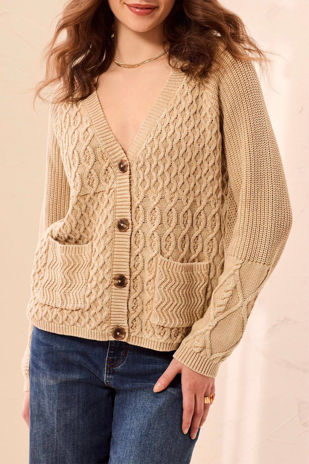 A woman wearing a beige cable knit cardigan and jeans standing in front of a beige background. 