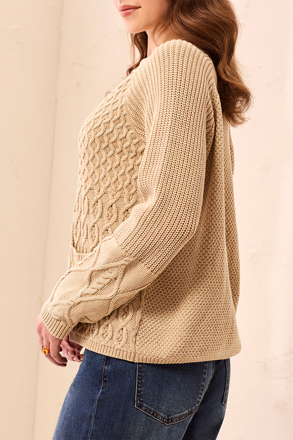 A woman shown from the side wearing a beige cable knit cardigan and jeans standing in front of a beige background. 