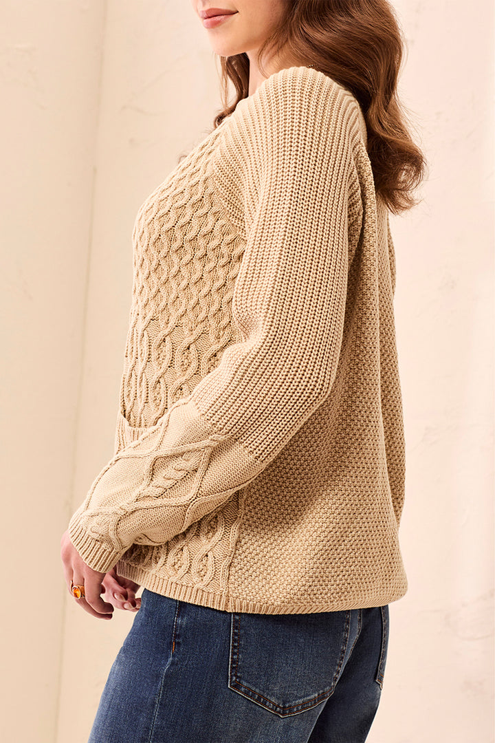 A woman shown from the side wearing a beige cable knit cardigan and jeans standing in front of a beige background. 