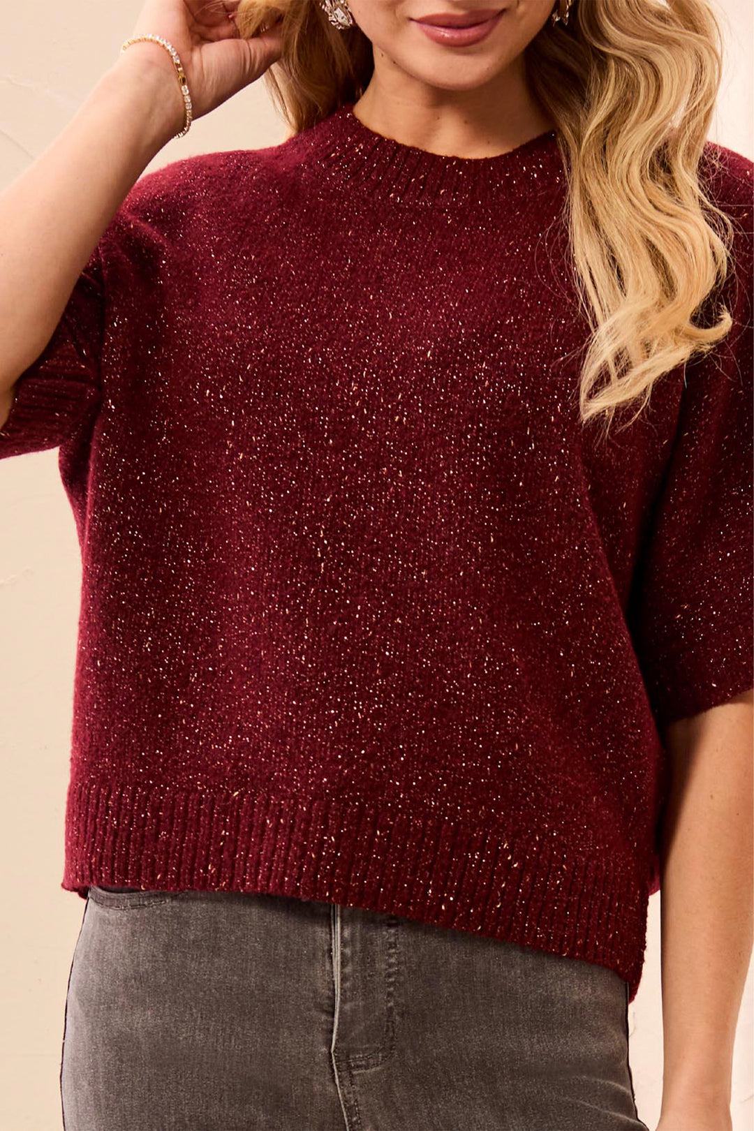 A woman wearing a deep read sparkly short sleeve boxy sweater standing in front of a beige background. 