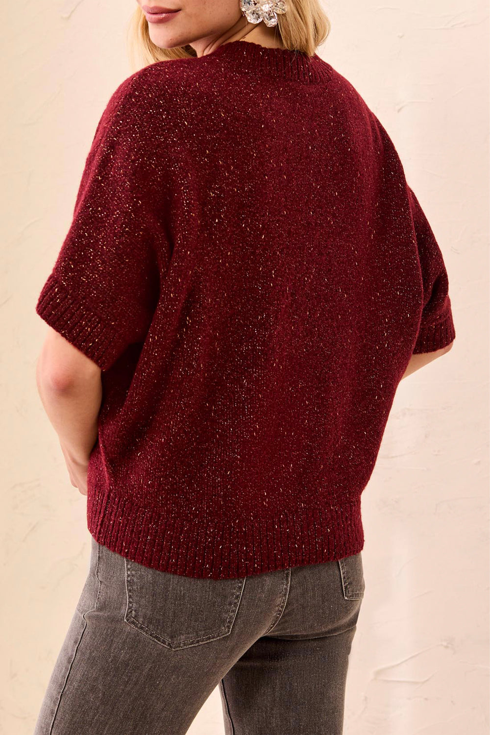 A woman shown from the back wearing a deep read sparkly short sleeve boxy sweater standing in front of a beige background. 