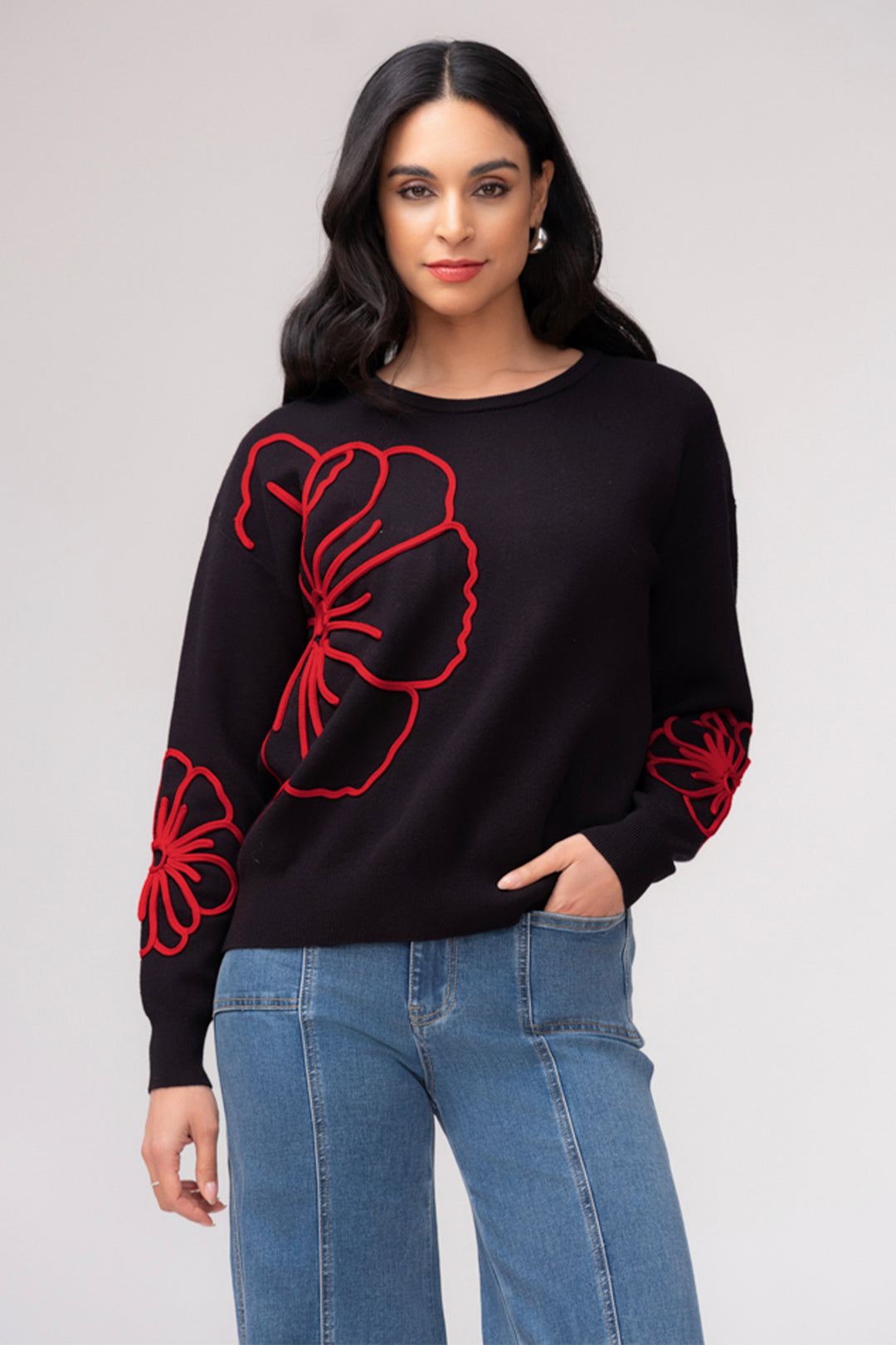 A woman wearing a black long sleeve sweater with large flower details. She is wearing jeans and standing in front of a light grey background. 