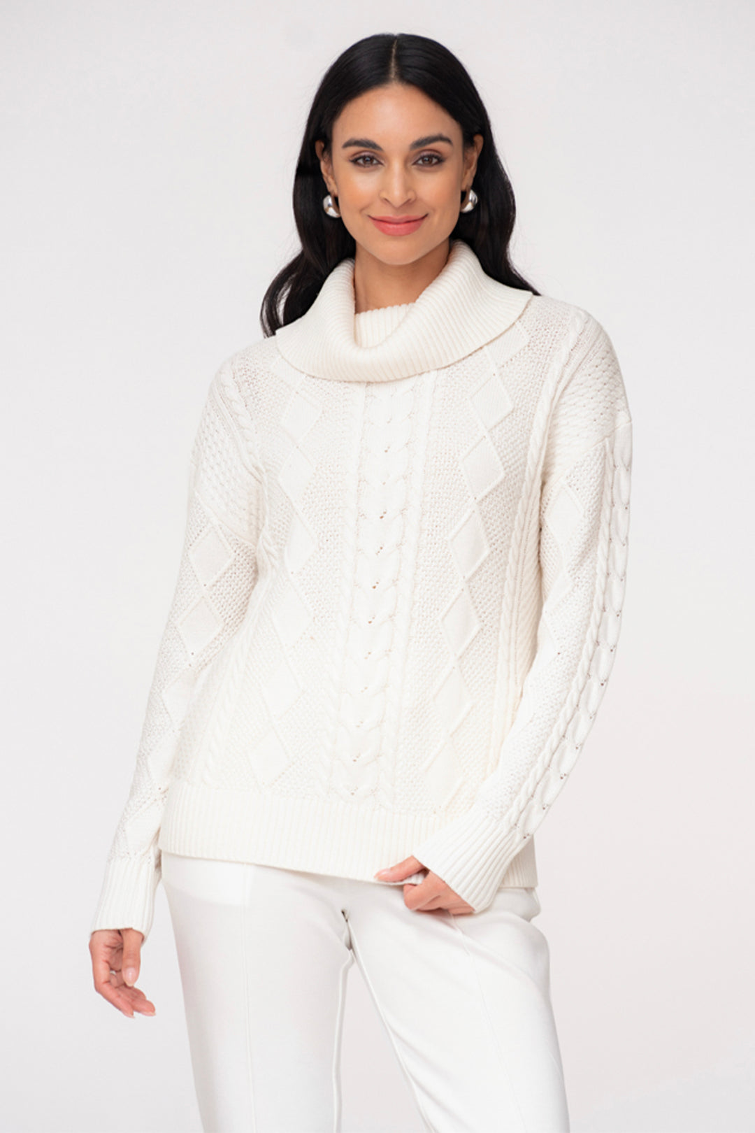 A woman wearing a white long sleeve cable knit sweater with a large cowl neck. She is wearing white pants and standing in front of a light grey background. 