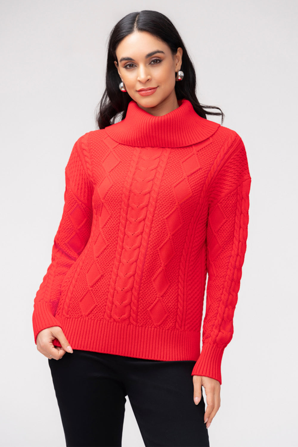 A woman wearing a red long sleeve cable knit sweater with a large cowl neck. She is wearing black pants and standing in front of a light grey background. 
