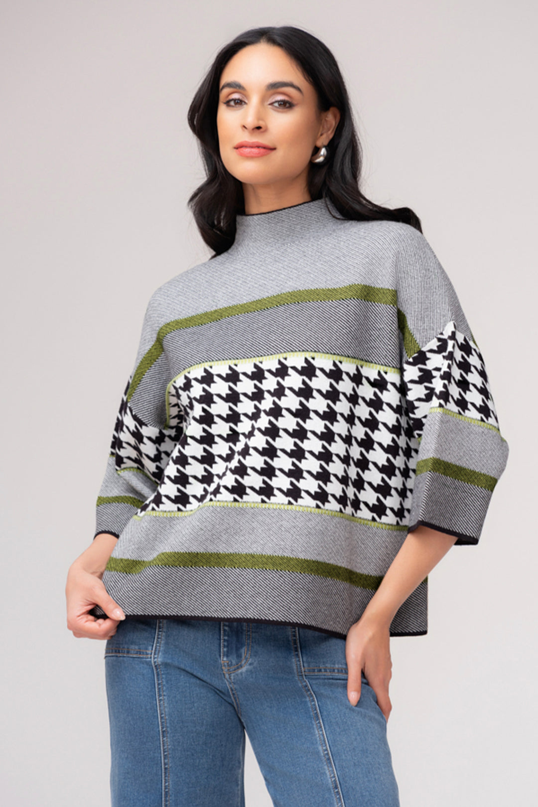 A woman wearing a grey, black, white and lime crop sleeve mock neck sweater. She is wearing jeans and standing in front of a light grey background. 