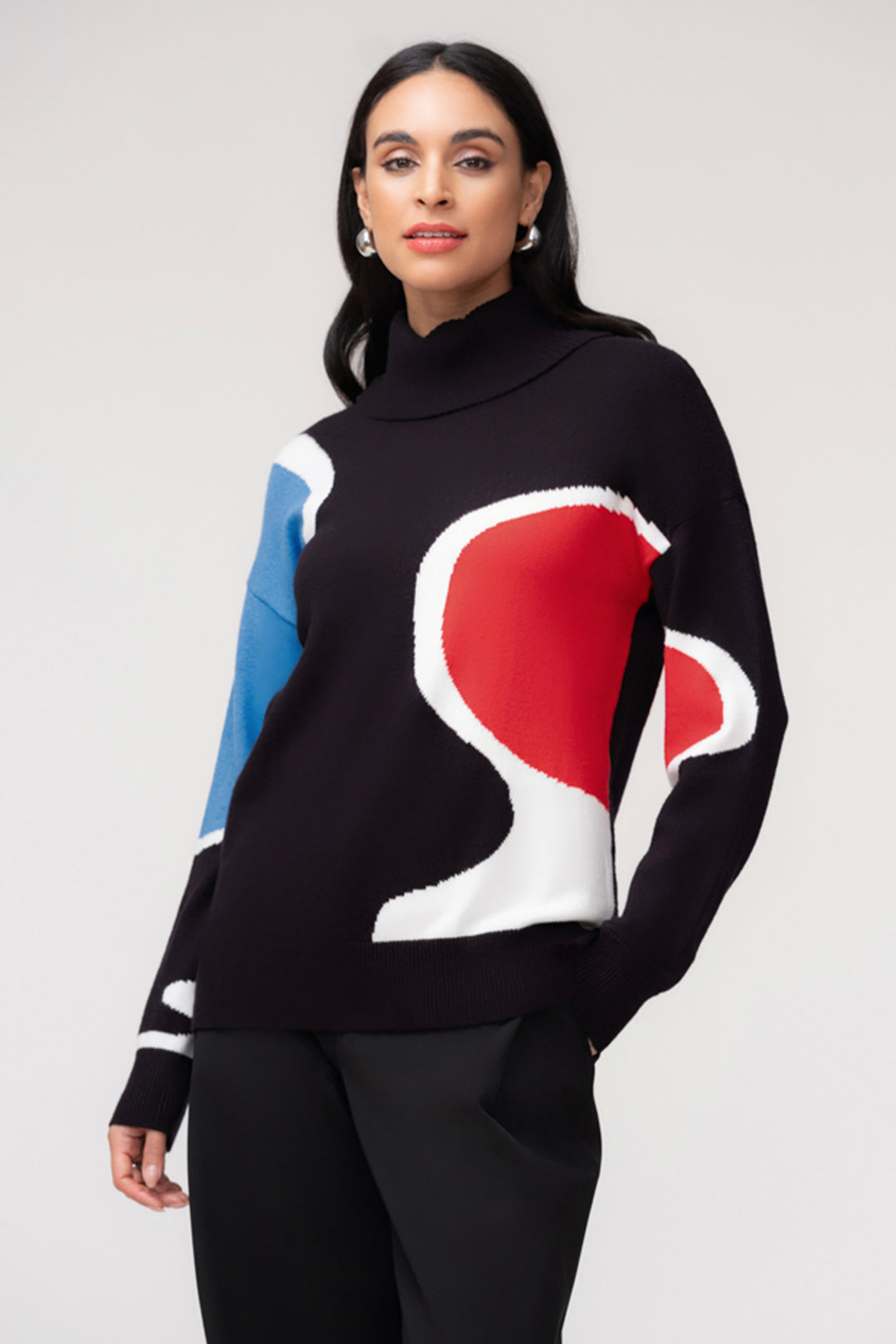 A woman wearing a black cowl neck sweater with a white, blue and red abstract print. She is wearing black pants and standing in front of a light grey background. 