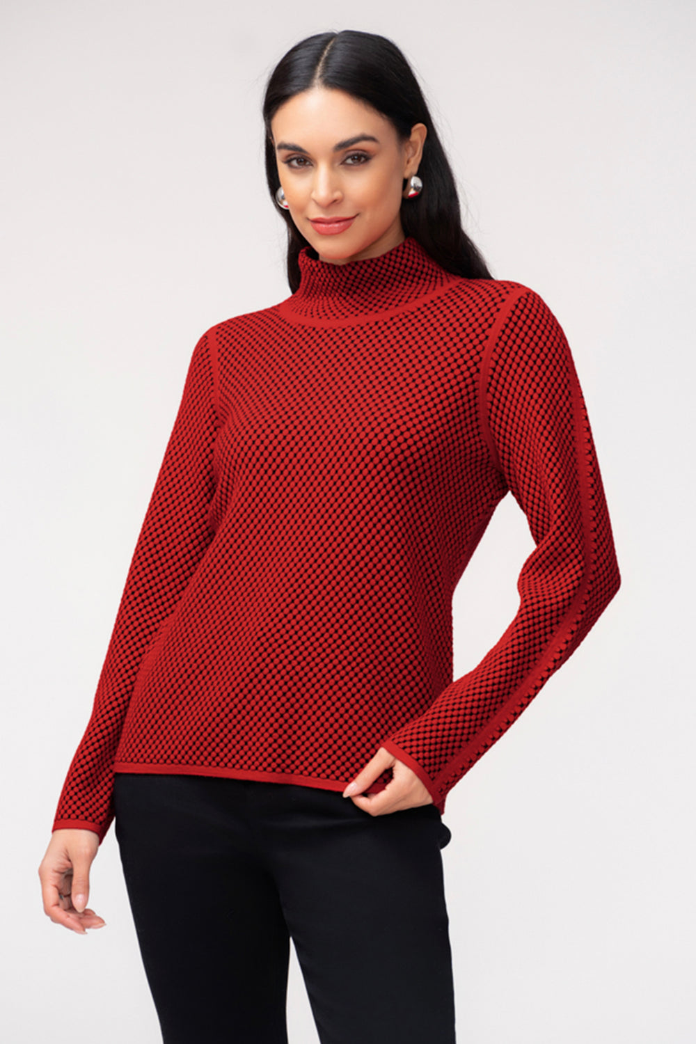 A woman wearing a red waffle print long sleeve sweater with a mock neckline. She is wearing black pants and standing in front of a light grey background. 