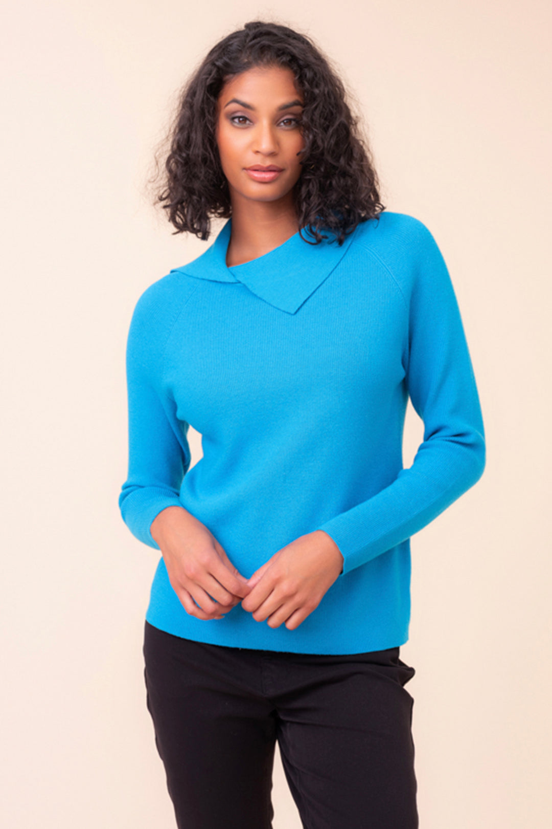 A woman wearing black pants and a bright teal pull over sweater with a split neck standing in front of a light beige background. 