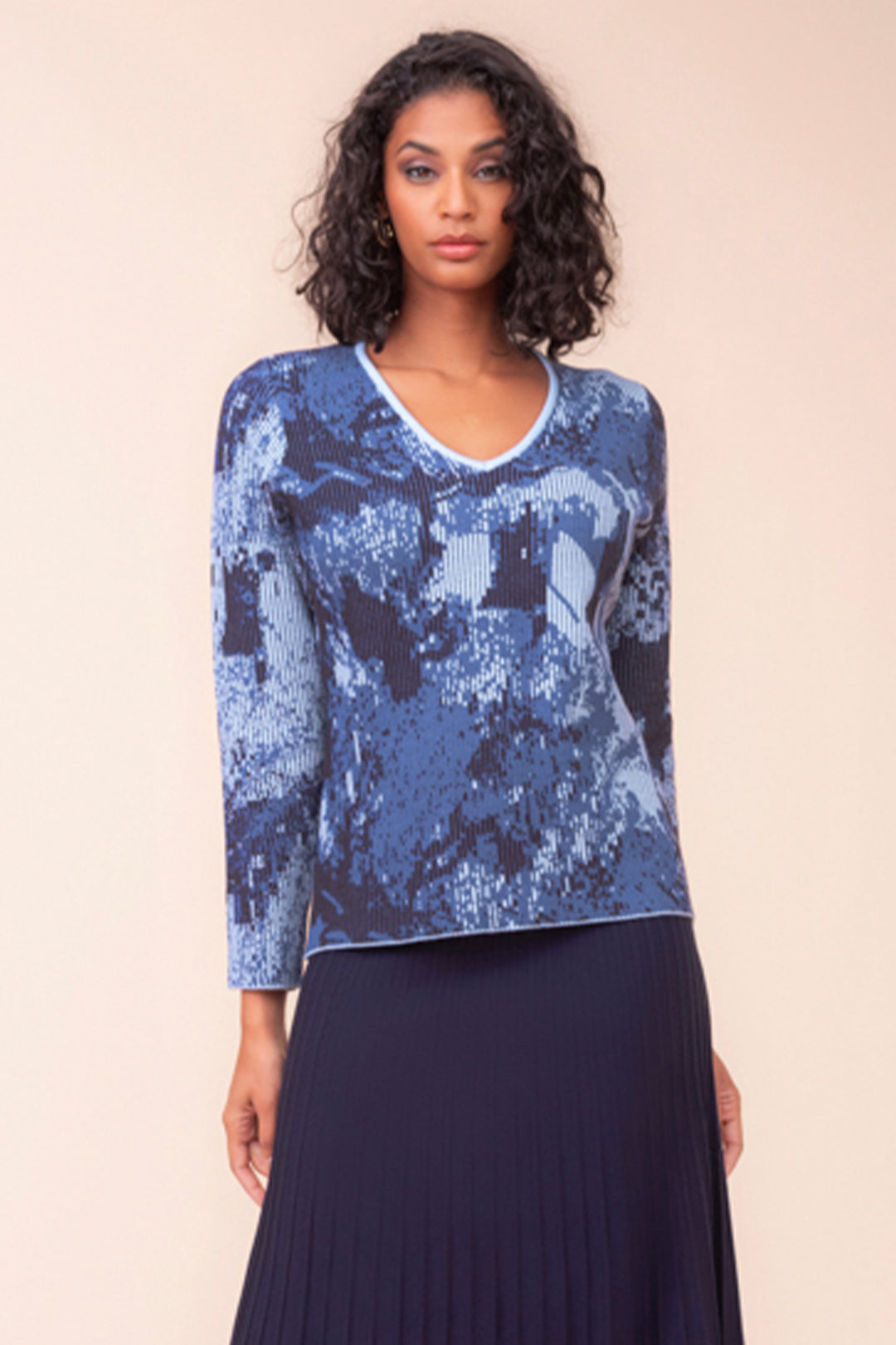 A woman wearing a blue and navy patterned long sleeve sweater and a navy pleated skirt. She is standing in front of a beige background. 