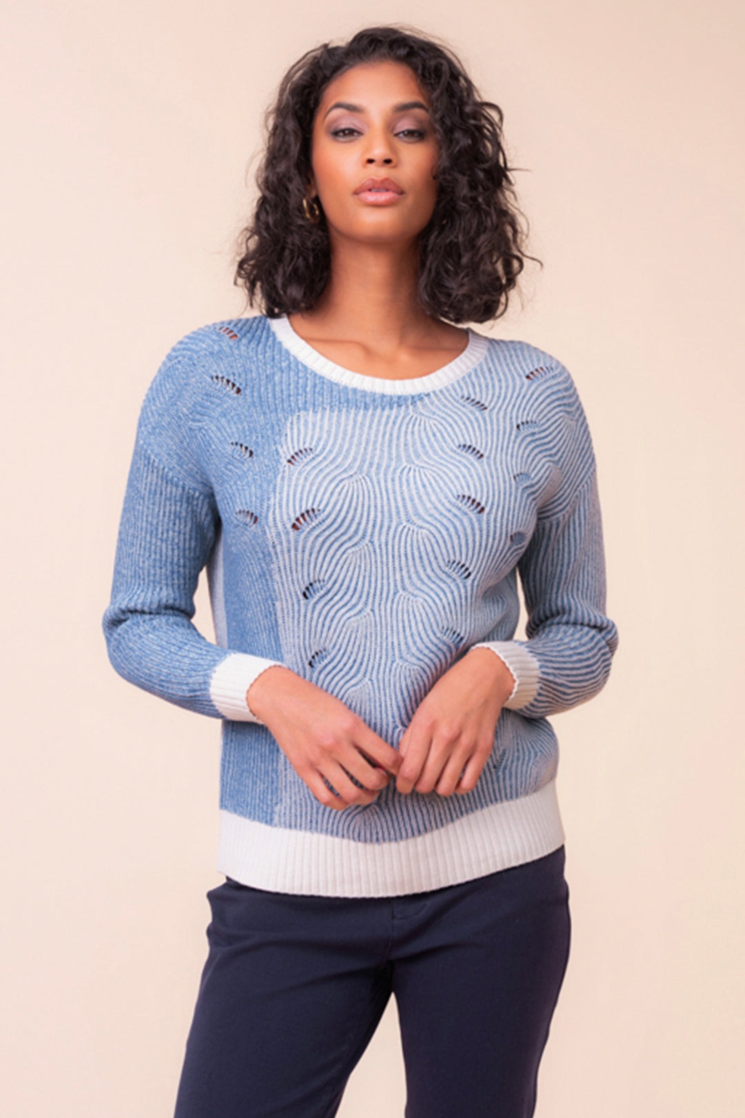 A woman wearing a blue long sleeve cable knit sweater standing in front of a beige background. 