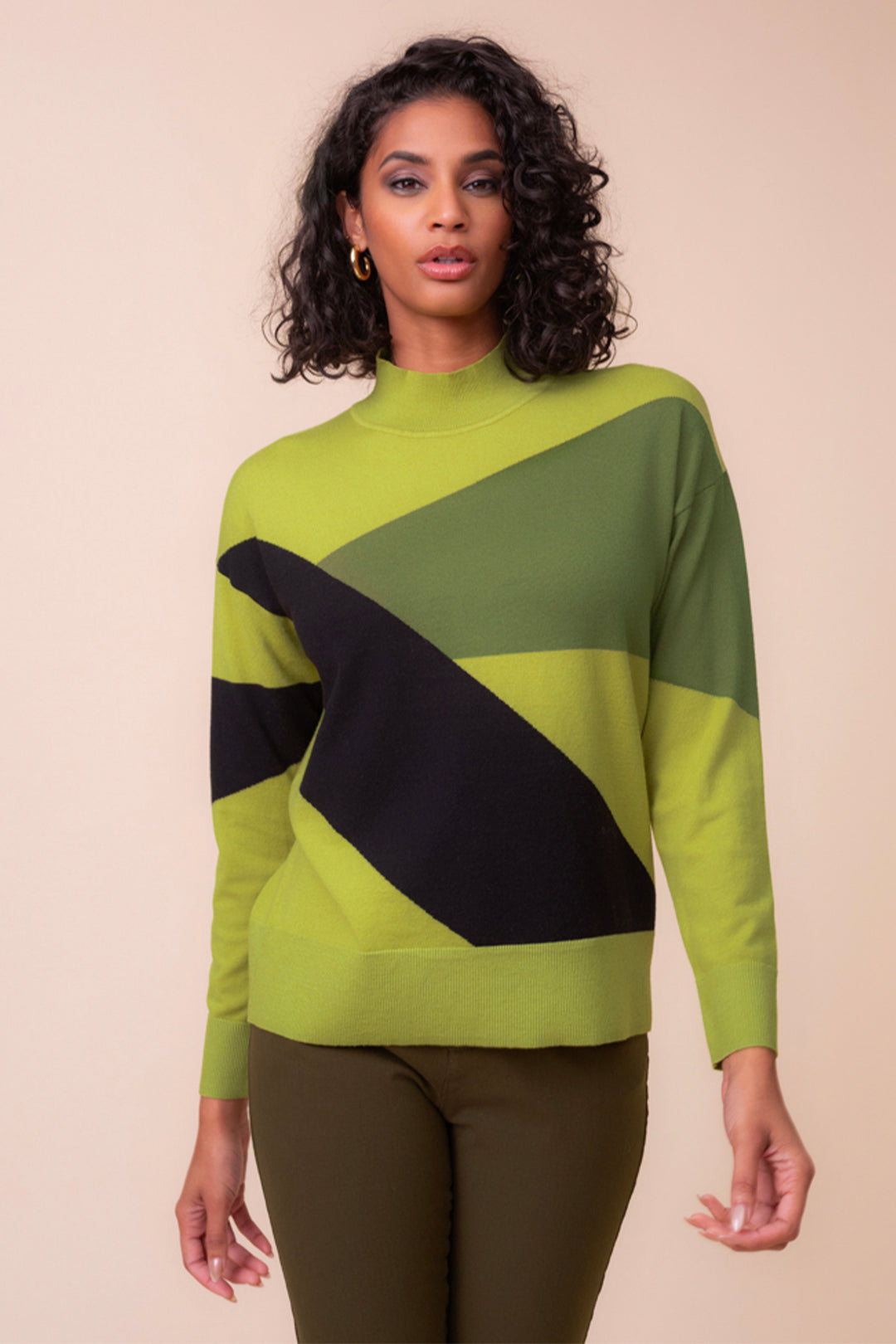 A woman wearing a lime green and black long sleeve sweater with brown pants standing in front of a light beige background. 