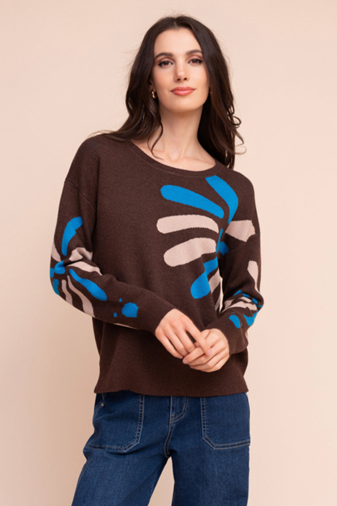 A woman wearing a brown long sleeve sweater with a large daisy-like print all over. She is standing in front of a beige background. 