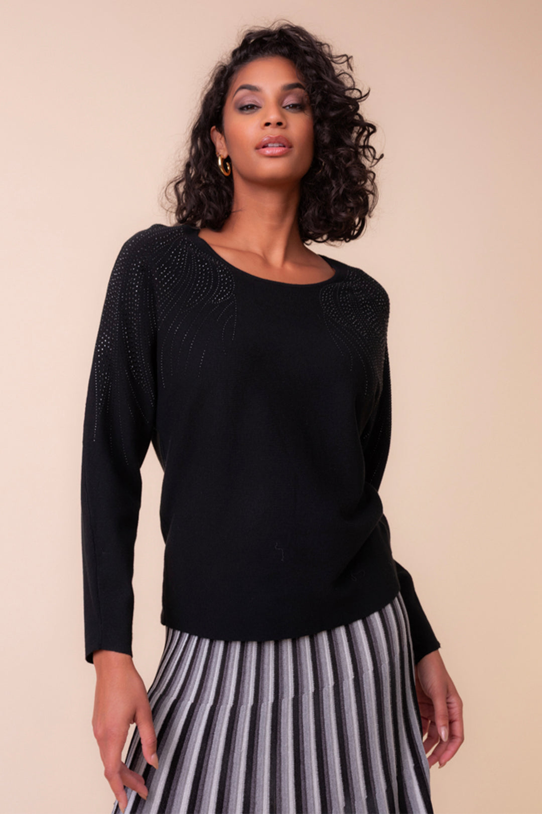  woman wearing a long sleeve boat neck sweater with rhinestone embellishments on the shoulders. She is standing in front of a light beige background. 