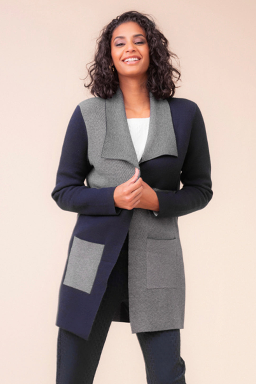 A woman wearing a navy and grey colourblock cardigan with large front pockets standing in front of a beige background. 