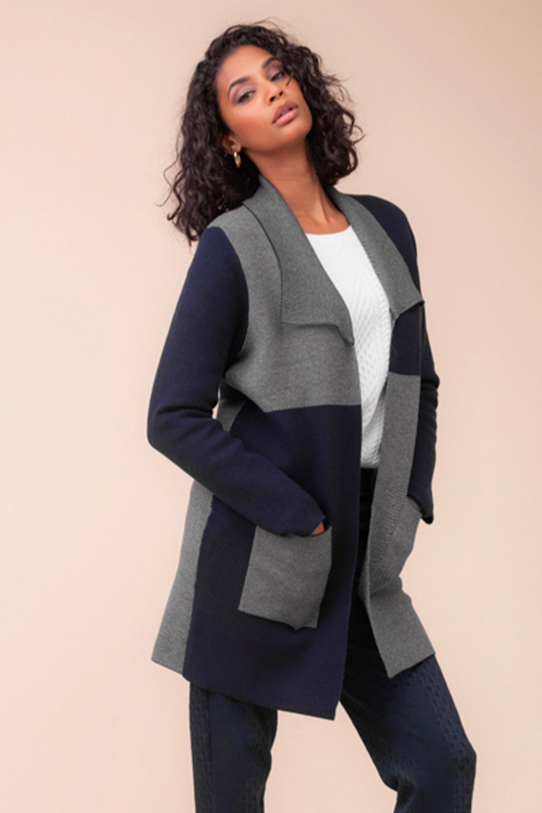 A woman wearing a navy and grey colourblock cardigan with large front pockets standing in front of a beige background. 