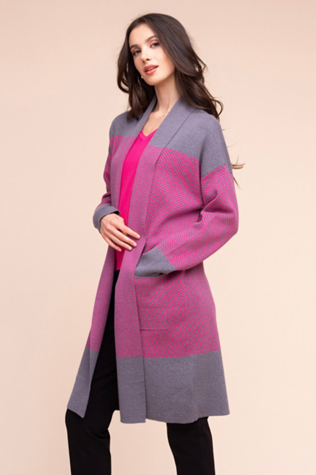 A woman wearing a long cardigan in grey in pink colour block pattern. She is standing in front of a light beige background. 