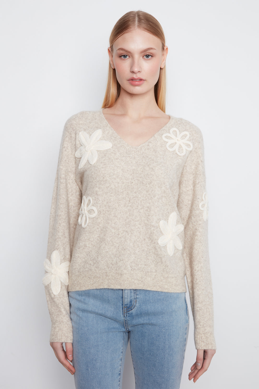 A woman wearing a long sleeve beige sweater with cream embroidered flowers on the front. She is wearing blue jeans and standing in front of a light grey wall. 