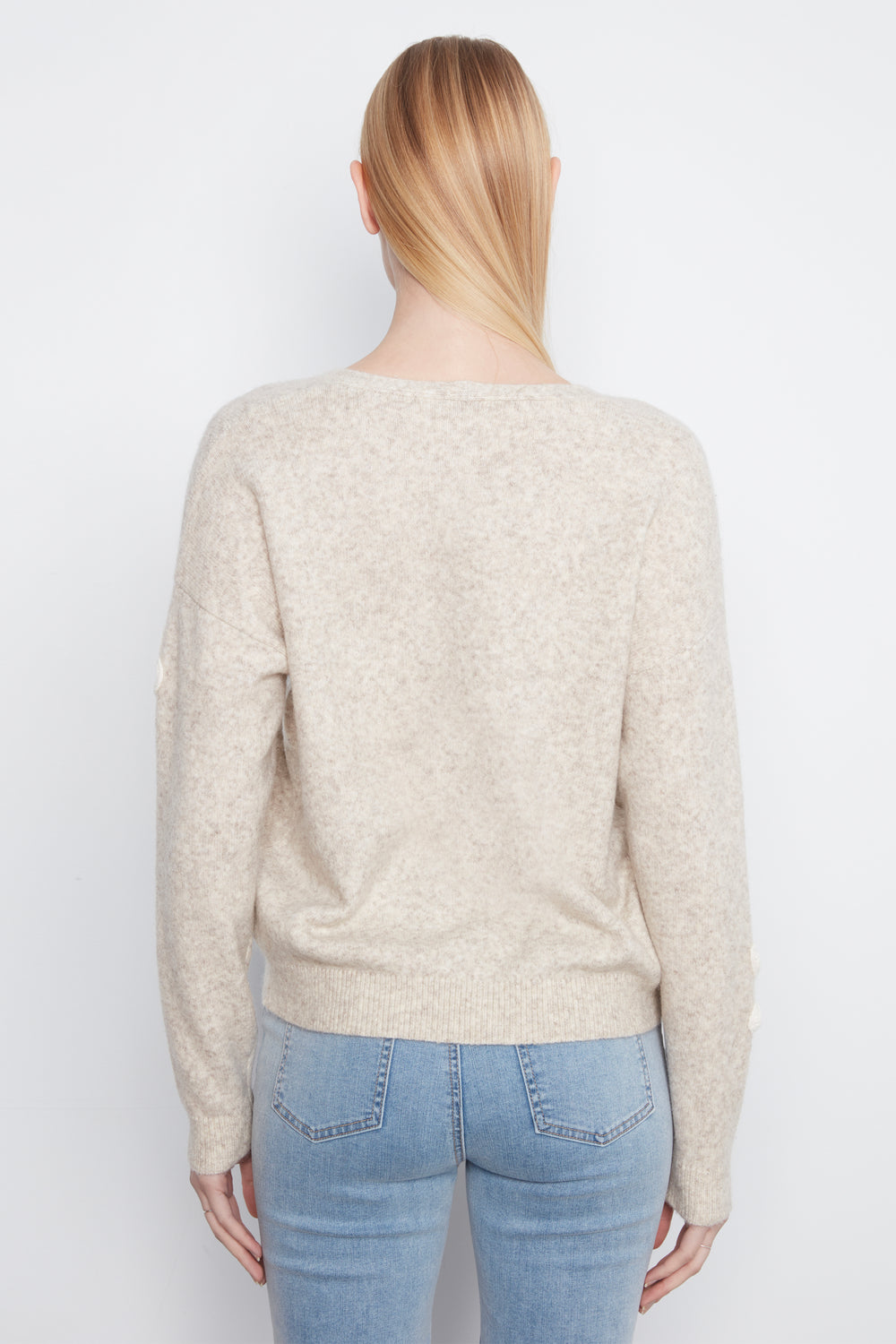 A woman shown from the back wearing a long sleeve beige sweater with cream embroidered flowers on the front. She is wearing blue jeans and standing in front of a light grey wall. 